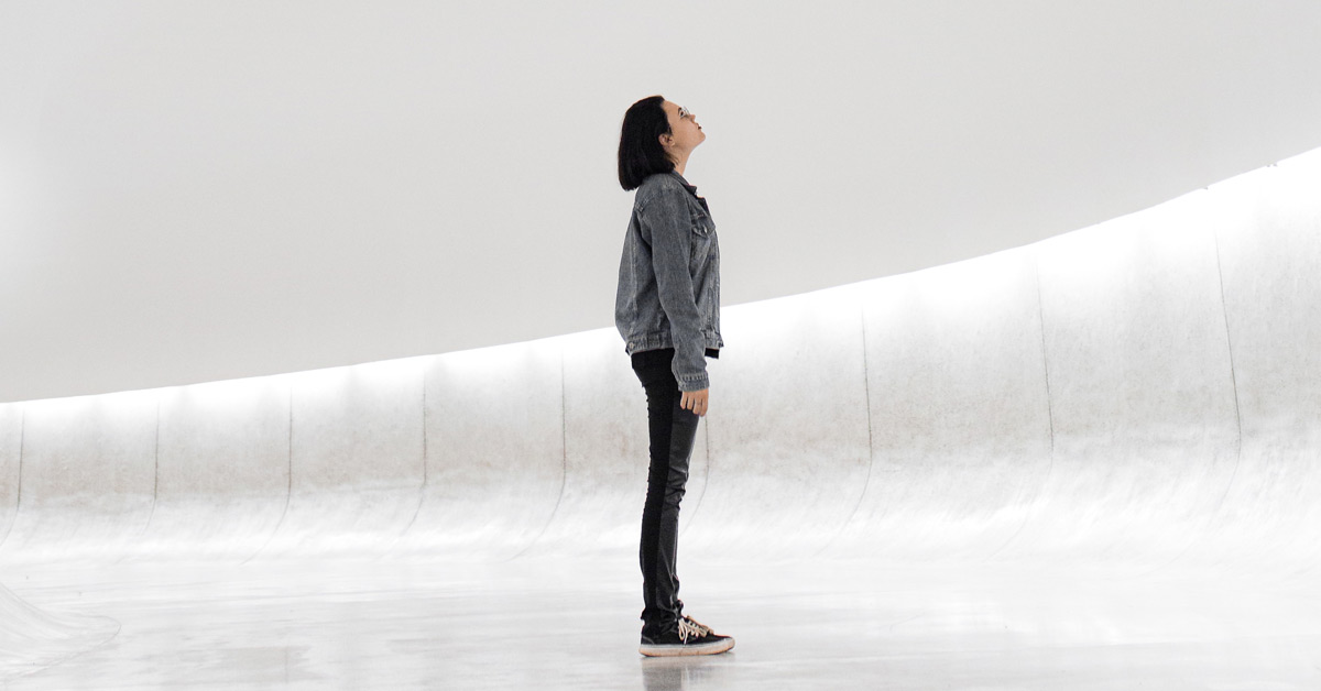Young woman in black jeans and gray denim jacket standing in futuristic space, calmly looking up.