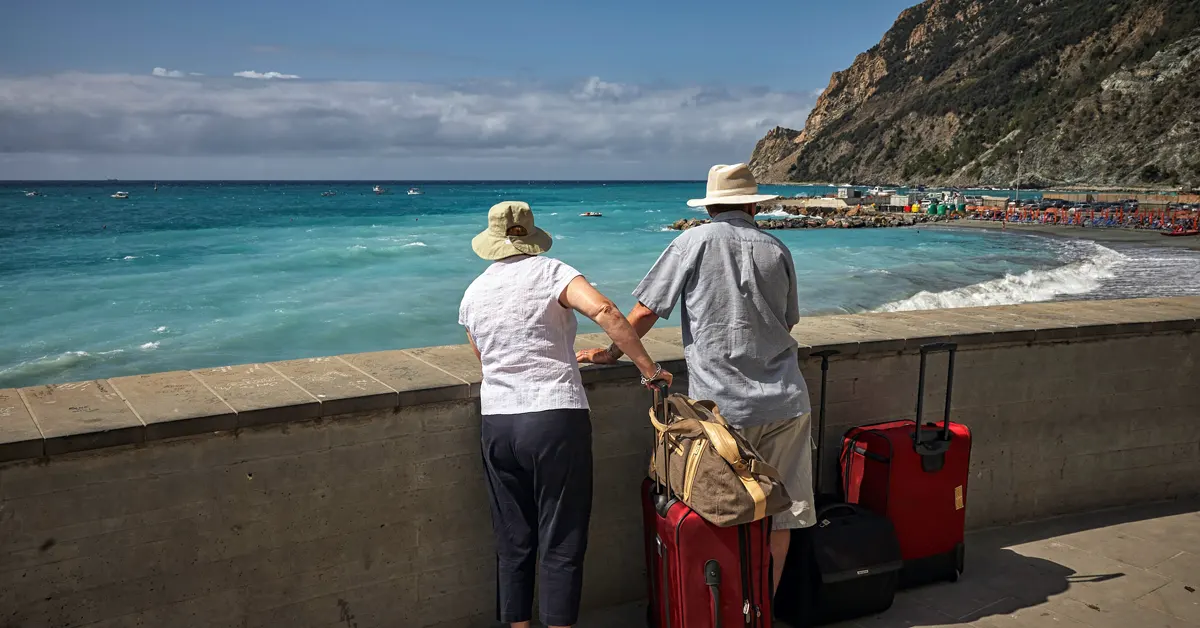 An elderly couple stands near the sea with their two red suitcases.