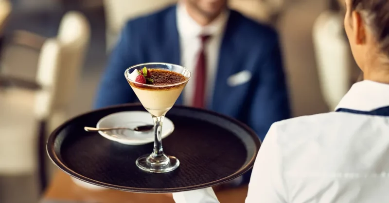 A hotel trainee waitress serving a hot coffee to a café terrace guest.